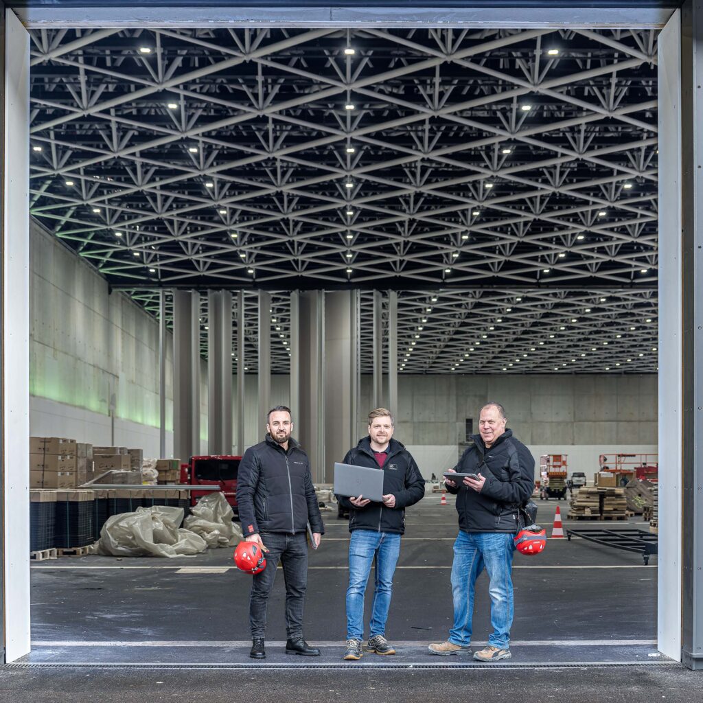 Drei Mitarbeitende der Hälg & Co. AG St. Gallen stehen vor den riesigen Toren der SGKB-Halle, welche sich noch im Bau befindet.