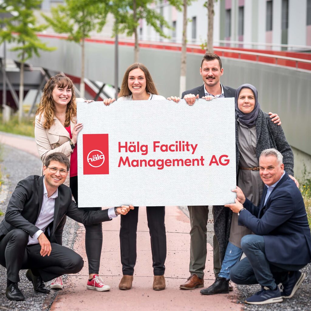 Fünf Mitarbeitende der Hälg Facility Management AG halten ein Schild mit dem Logo der HFM AG.